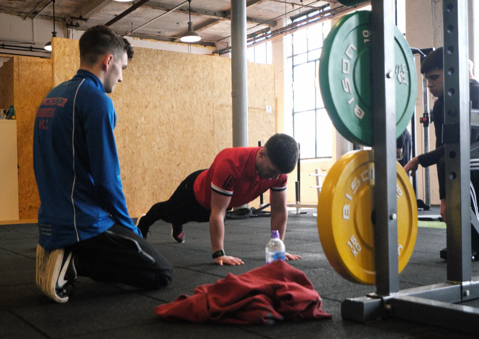 A tutor kneeling beside a learner on the gym floor, coaching a press-up